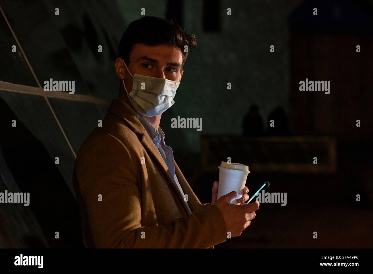 Junger Geschäftsmann mit Gesichtsmaske, Kaffeetasse und Telefon in der Abenddämmerung Stockfoto