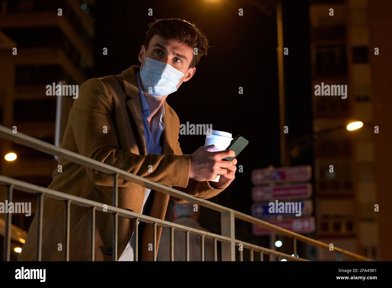 Junger Geschäftsmann mit Gesichtsmaske, Kaffeetasse und Telefon in der Abenddämmerung Stockfoto