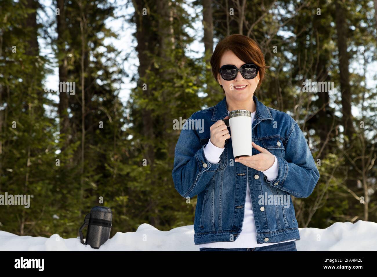 Nachbau einer Frau, die eine Sonnenbrille trägt und einen weißen Reisebecher in einem verschneiten Wald hält. Leere Reise-Kaffeebecher mit Griffschablone, Modell mockup Stockfoto