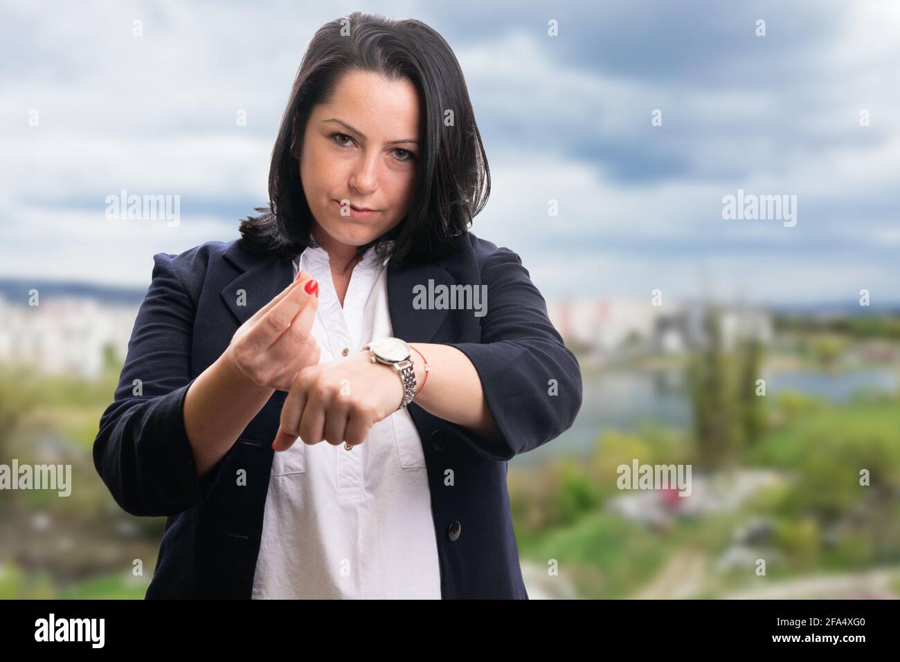 Geschäftsfrau mit ernstem Ausdruck in elegantem, legerem Büroanzug Zeit bedeutet Geldwert Geste zeigt Armbanduhr auf Stadt und Natur-Bac Stockfoto