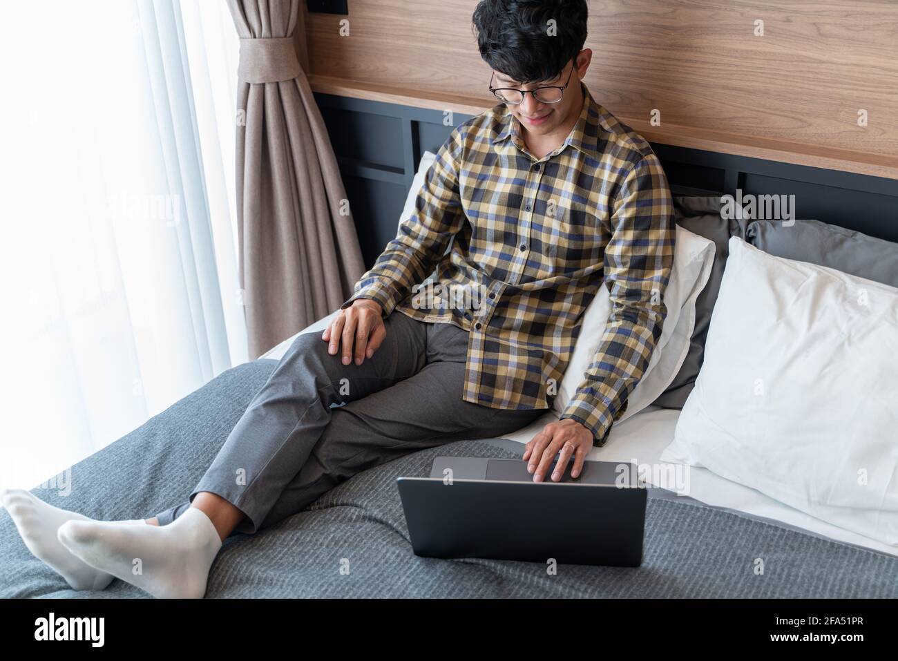 Arbeiten Sie von zu Hause aus, ein Mann mit kariertem Hemd, der eine Augenbrille trägt, liegt auf dem Bett, während er den ganzen Körper am Computer benutzt. Stockfoto