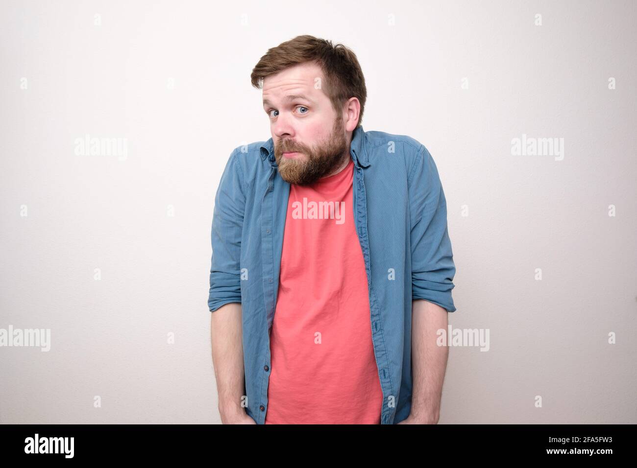 Verwirrender bärtiger Mann in legerer Kleidung sieht überlegt und überrascht aus, wirft Hände in die Taschen und hebt Schultern. Weißer Hintergrund. Stockfoto