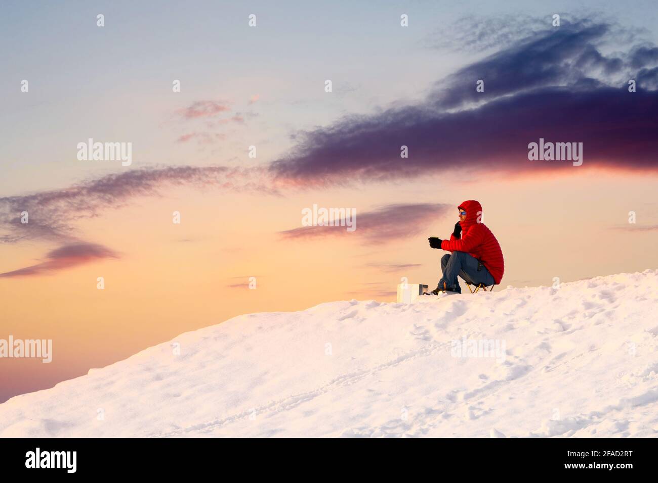 Wanderer Bergsteiger in leuchtend rotem Mantel und Sonnenbrille saßen und beobachteten den unglaublichen orangefarbenen Sonnenaufgang über der schneebedeckten Bergspitze. High-Hill-Kaffee. Stockfoto