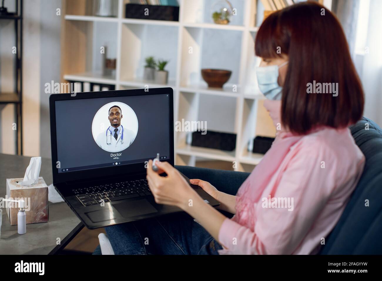 Verschwommenes Bild einer jungen kranken Frau in einer Gesichtsmaske, die auf dem Sofa sitzt und auf den Thermometerbildschirm schaut, der ihre Körpertemperatur überprüft, während sie einen Videoanruf über den pc mit ihrem Arzt führt. Konzentrieren Sie sich auf Notebooks. Stockfoto