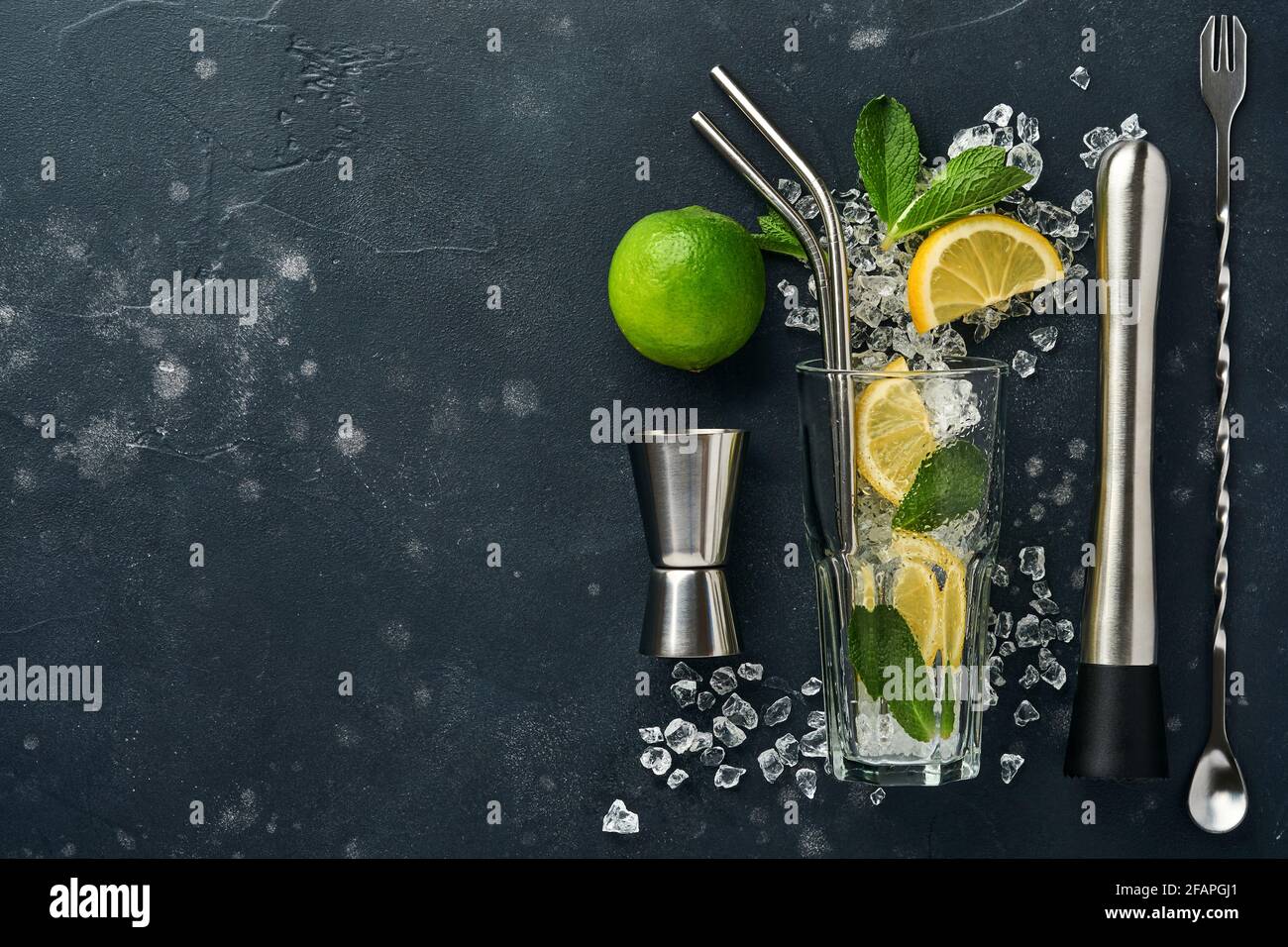 Mojito oder Lemonade Cocktail im Longdrinkglas mit Eiswürfeln, Minze und Limette auf schwarzem Steingrund mit Shaker und Becher. Menüleiste. Platz für Text Stockfoto