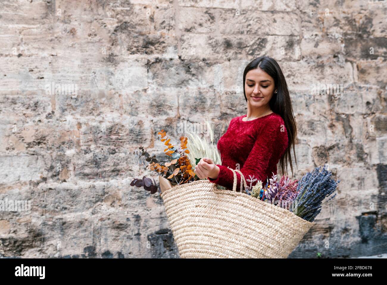 Schöne Frau, die Strohbeutel mit Blumen trägt, während sie steht Wand Stockfoto