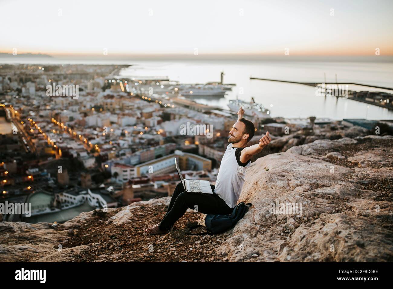 Sorgloser Mann mit Laptop, der während des Sonnenaufgangs auf dem Berg sitzt Stockfoto