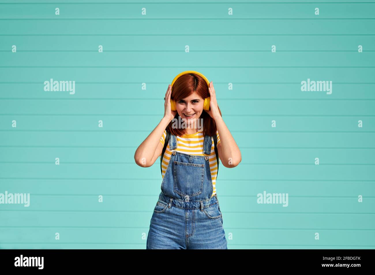 Junge Frau, die im Stehen Musik über kabellose Kopfhörer hört Vorderseite der türkisblauen Wand Stockfoto