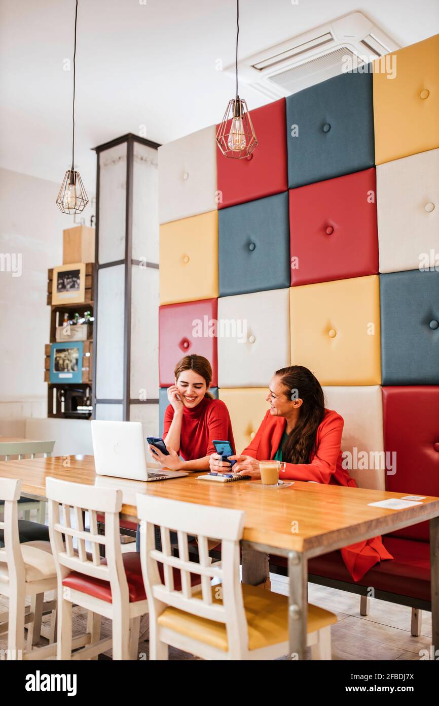 Lächelnde freiberufliche Arbeitnehmerinnen, die beim Sitzen Mobiltelefone halten Tisch im Café Stockfoto