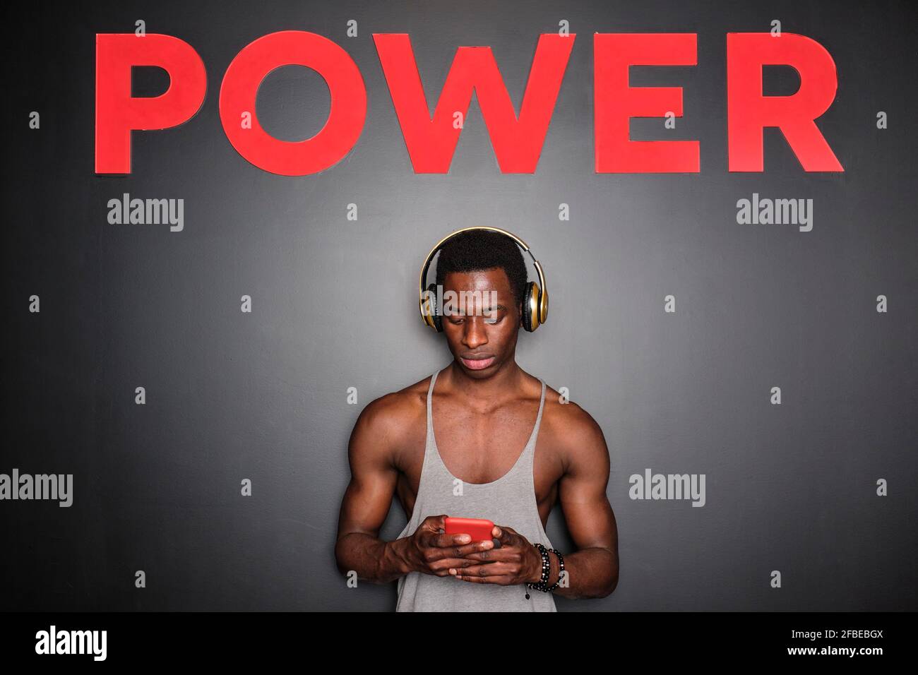 Junger afrikanischer Mann mit Kopfhörern und Smartphone unter Strom Text an der Wand im Fitnessstudio Stockfoto