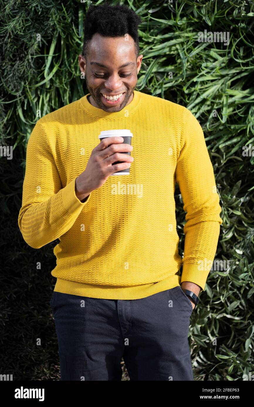 Lächelnder junger Mann, der vor dem Hotel die Einweg-Kaffeetasse anschaut Von üppigem Laub Stockfoto