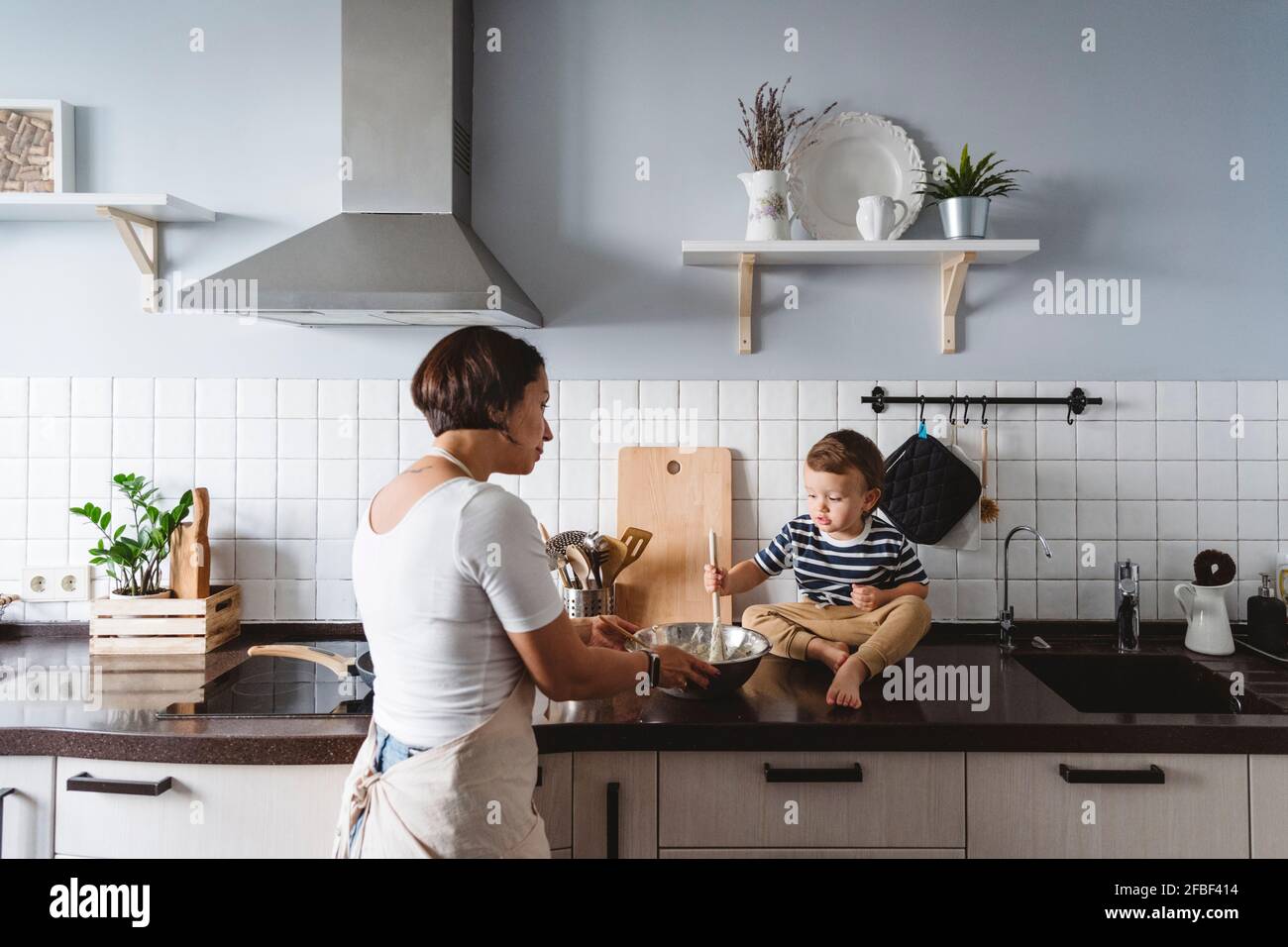 Mutter sieht sich ein niedliches Kind an, das mit einer Schüssel auf dem Tisch spielt Küchentisch zu Hause Stockfoto