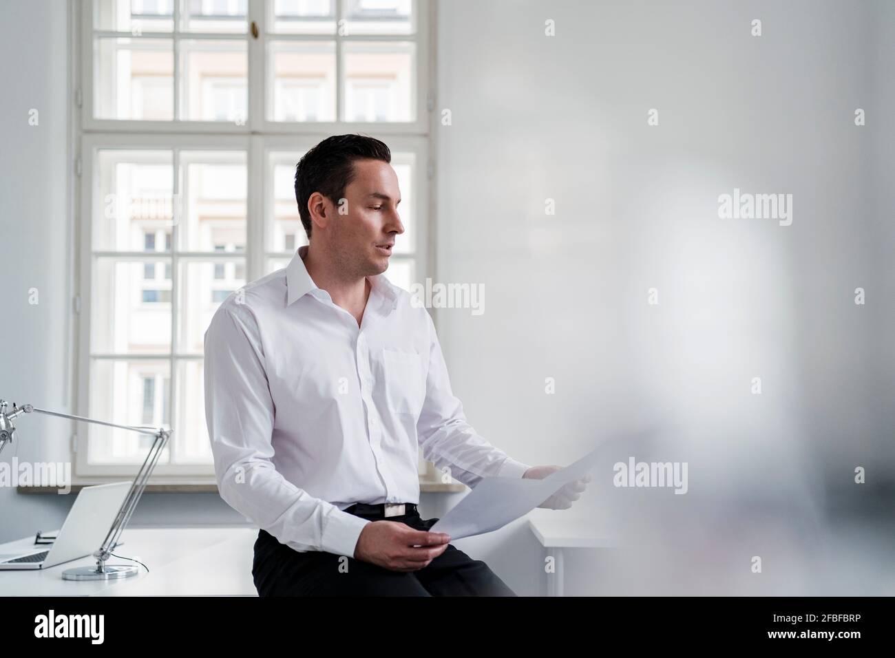Männlicher Unternehmer prüft Dokument, während er sich am Arbeitsplatz auf den Schreibtisch lehnt Stockfoto