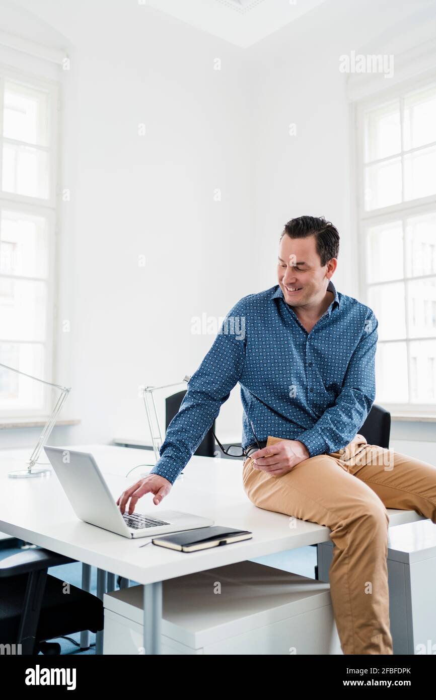 Männlicher Profi sitzt auf dem Schreibtisch, während er am Arbeitsplatz einen Laptop benutzt Stockfoto