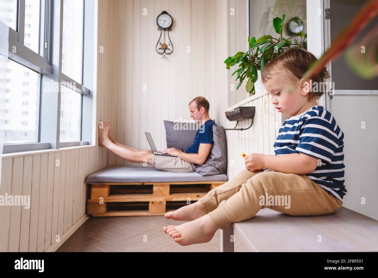 Spielerisches Kind sitzt auf dem Tisch, während der Vater am Laptop arbeitet Im Home Office Stockfoto