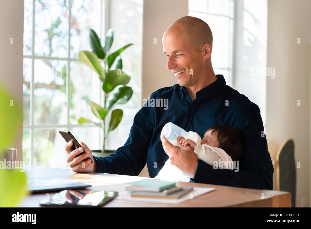 Vater füttert Baby Mädchen Milch, während er an smart arbeitet Telefon im Heimbüro Stockfoto