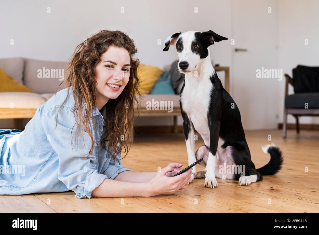 Hübsche Frau, die mit einem Smartphone auf Holzboden liegt Hund sitzt neben ihr beide Blick in die Kamera Stockfoto