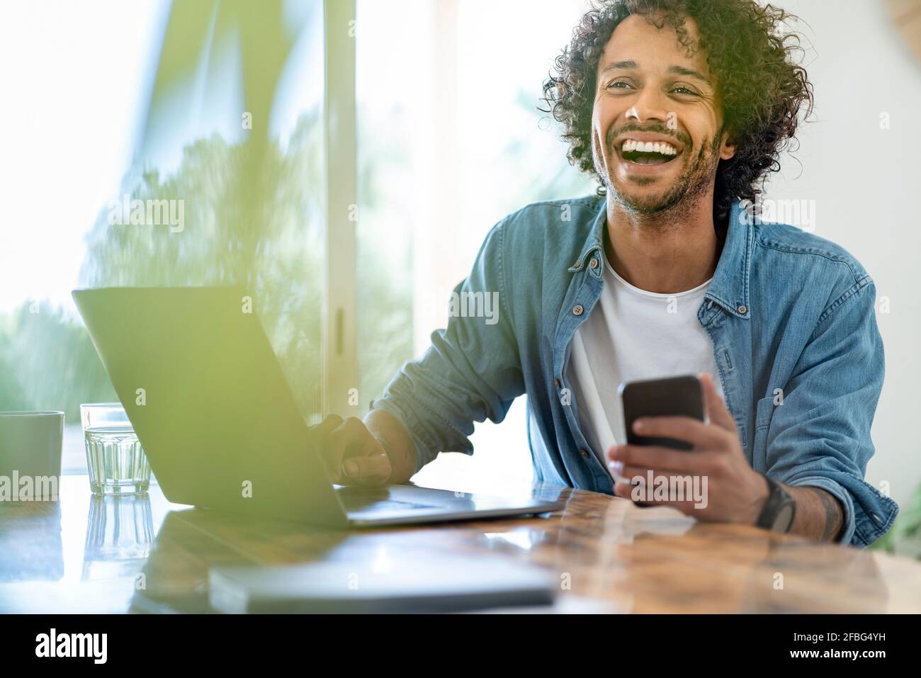 Fröhlicher Geschäftsmann mit Smartphone, während er mit einem Laptop sitzt Zu Hause Stockfoto