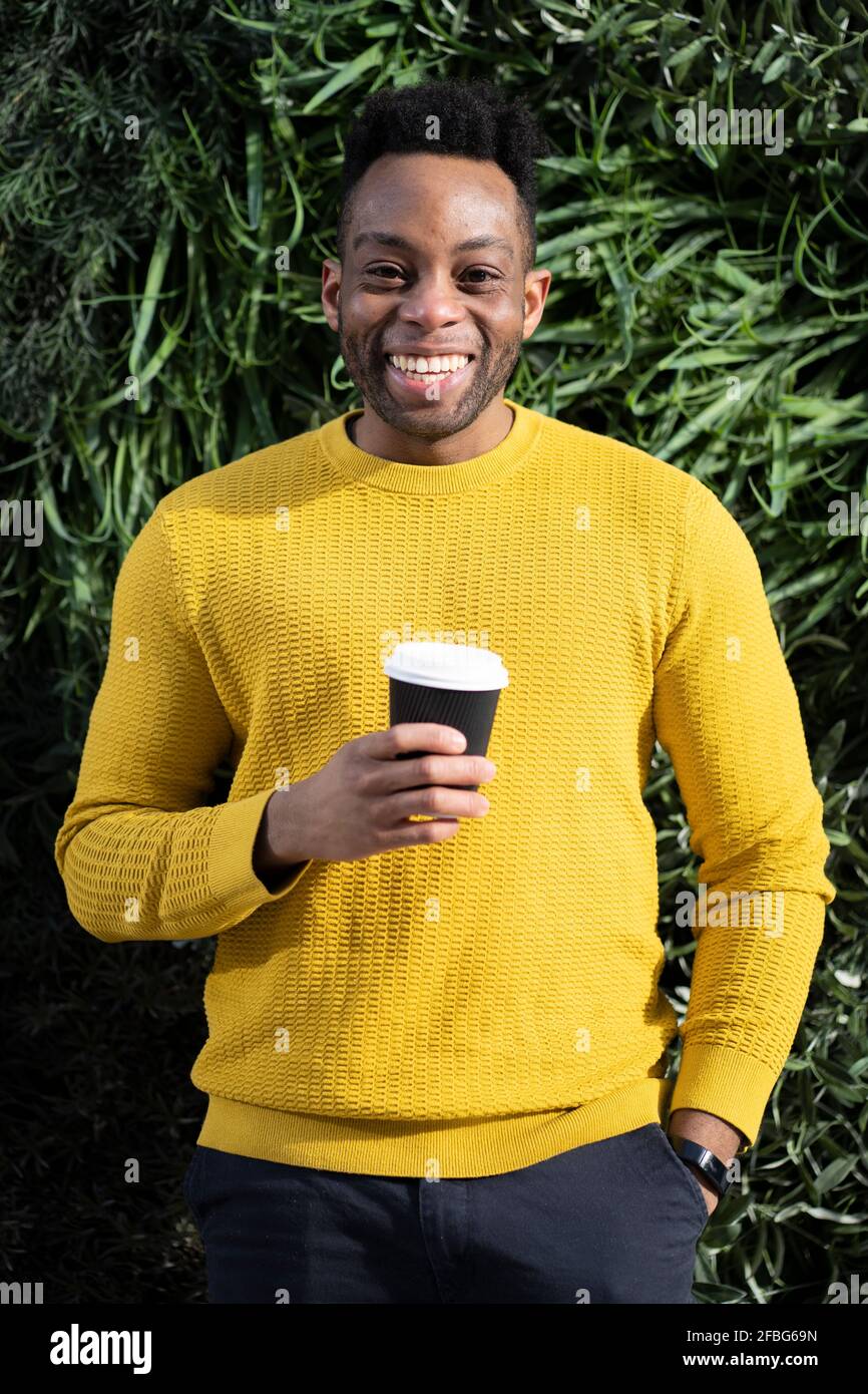 Glücklicher Mann mit der Hand in der Tasche, der die Einweg-Kaffeetasse hält Vor üppigem Laub Stockfoto