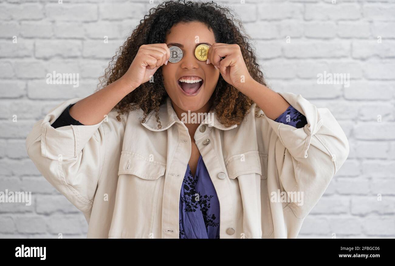 Aufgeregte, lockige Frau, die vor der Wand die Augen mit Gold- und silberfarbenen Bitcoins bedeckt Stockfoto