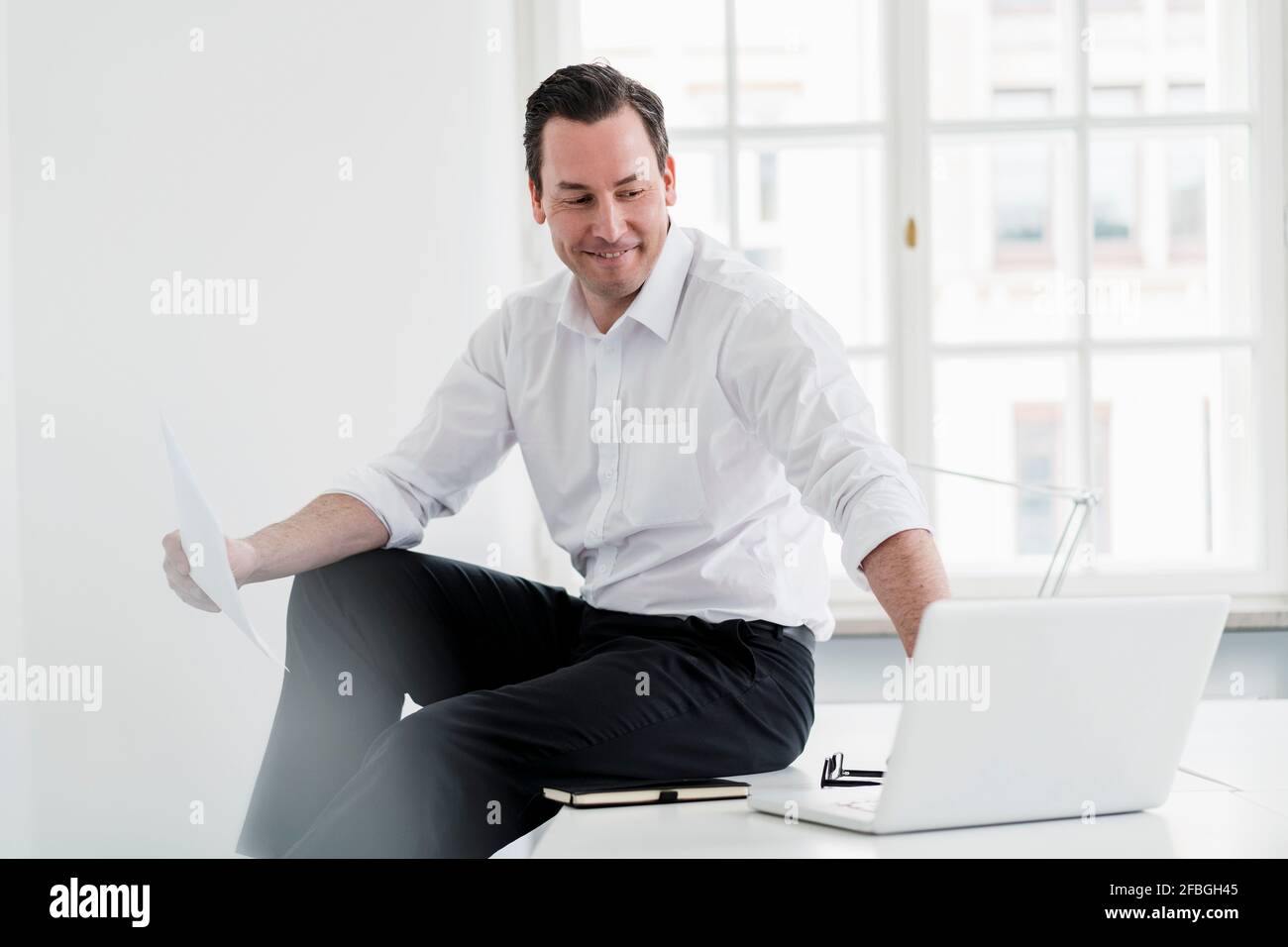 Männlicher Profi, der am Arbeitsplatz einen Laptop benutzt, während er auf dem Schreibtisch sitzt Stockfoto