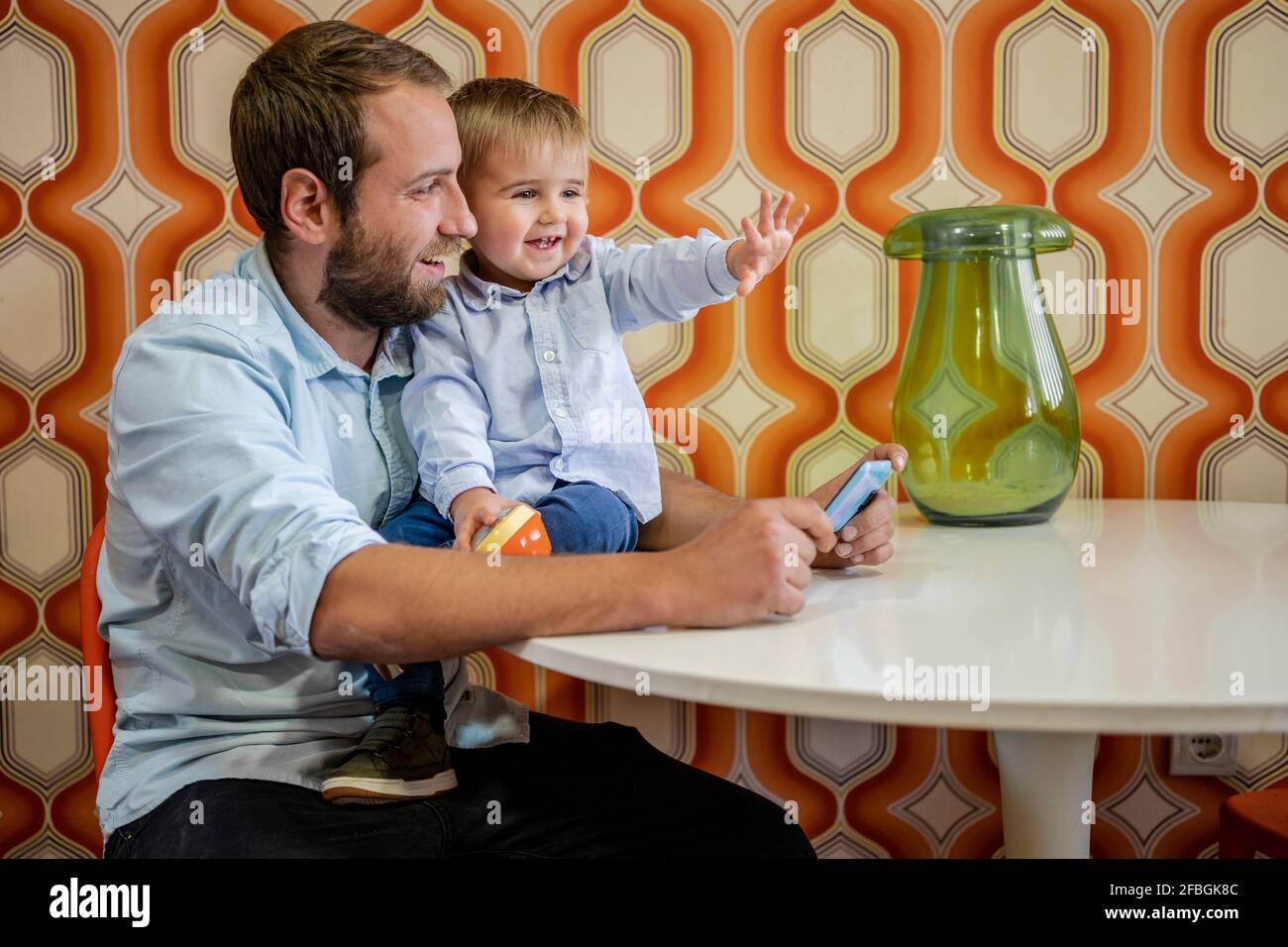 Lächelnder Sohn gestikuliert, während er vom Vater auf dem Tisch sitzt Zu Hause Stockfoto