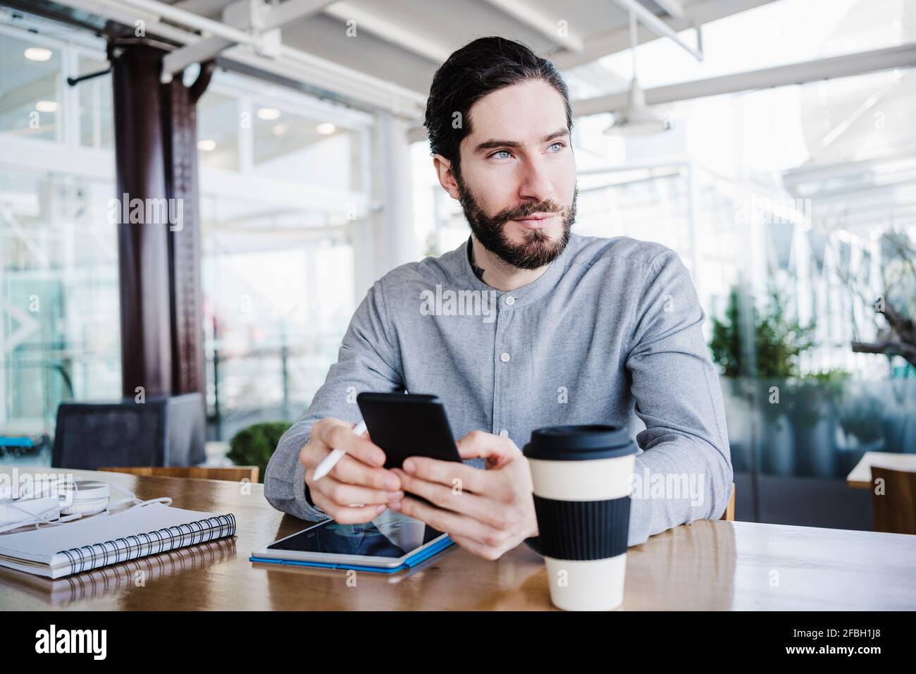Kontemplativer männlicher Unternehmer, der wegschaut, während er mit drahtlosen Technologien sitzt Am Arbeitsplatz Stockfoto