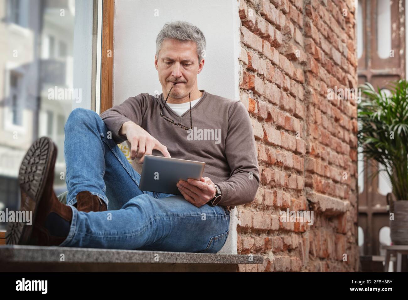 Mann, der ein digitales Tablet benutzt, während er die Brille im Mund hält Zu Hause Stockfoto