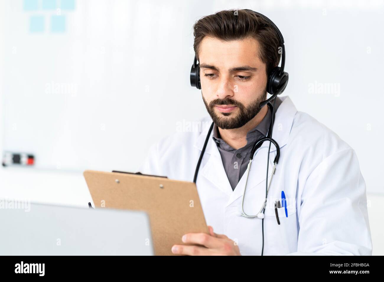 Hübscher männlicher Arzt, der Kopfhörer trägt, während er Bericht auf der Zwischenablage liest Im Krankenhaus Stockfoto