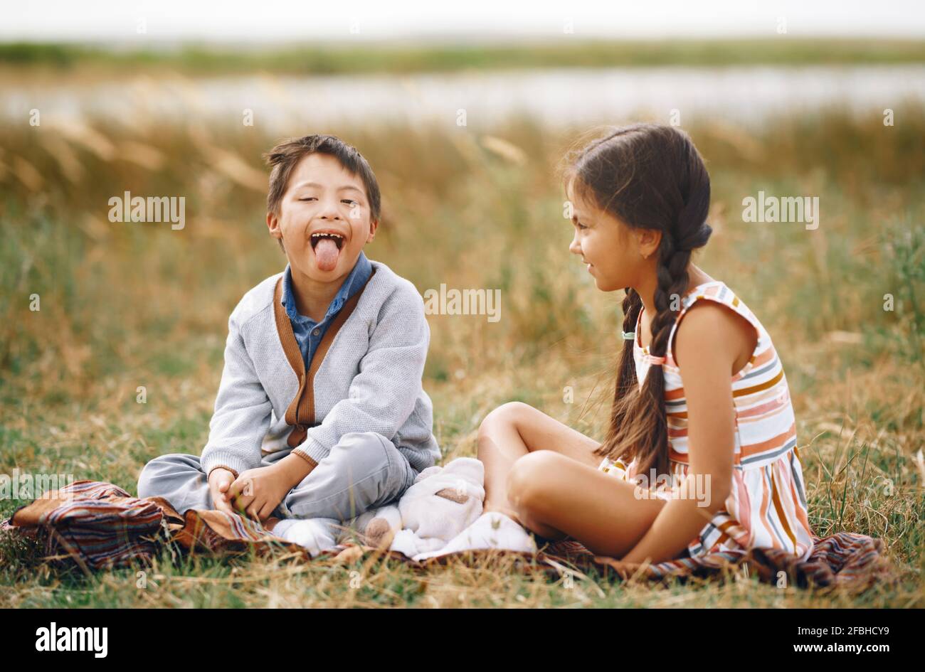 Mädchen, das Bruder mit Down-Syndrom betrachtet, der aus der Zunge ragt, während er auf dem Gras sitzt Stockfoto