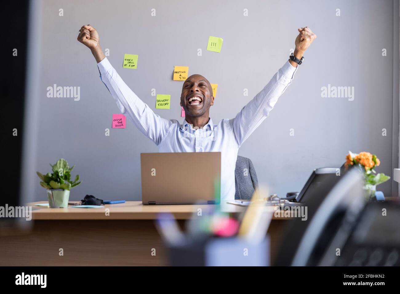 Fröhlicher Geschäftsmann mit erhobenen Armen, während er mit einem Laptop sitzt Büro Stockfoto