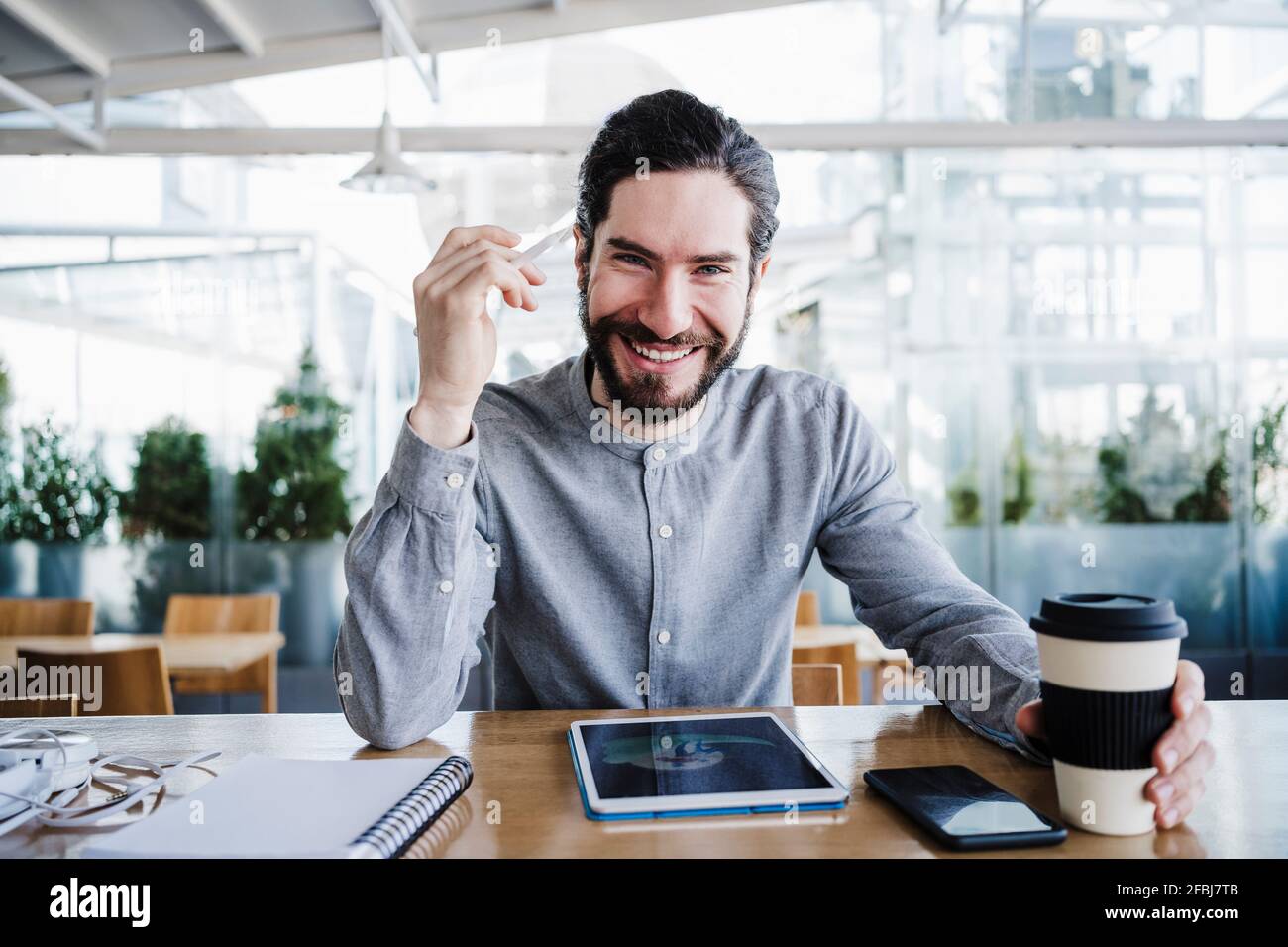 Lächelnder männlicher Freiberufler mit wiederverwendbarer Kaffeetasse durch drahtlose Technologien Im Büro Stockfoto