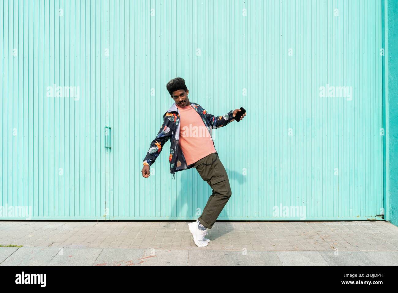 Ekstatischer Mann hält Smartphone, während er vor ihm tanzt Türkisfarbene Wand Stockfoto