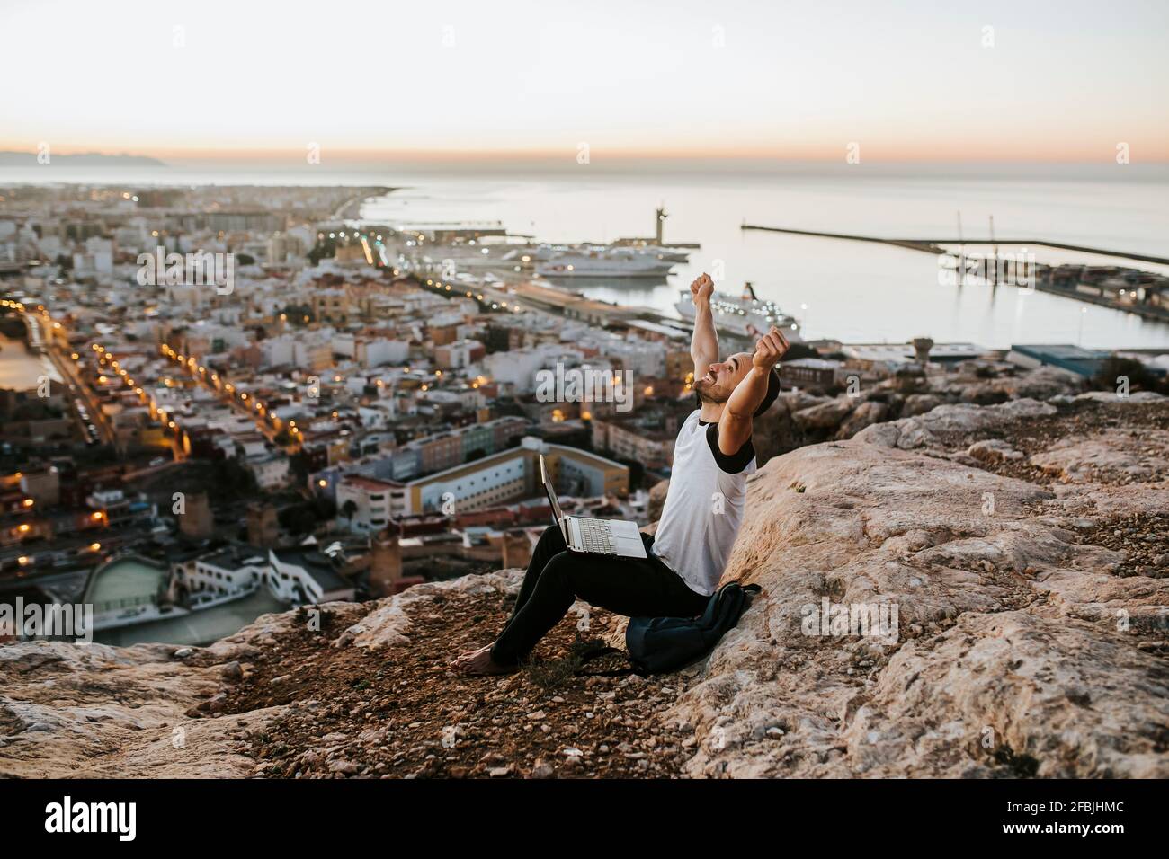 Unbeschwerter Mann mit ausgestreckten Armen, der während des Sonnenaufgangs auf dem Berg sitzt Stockfoto