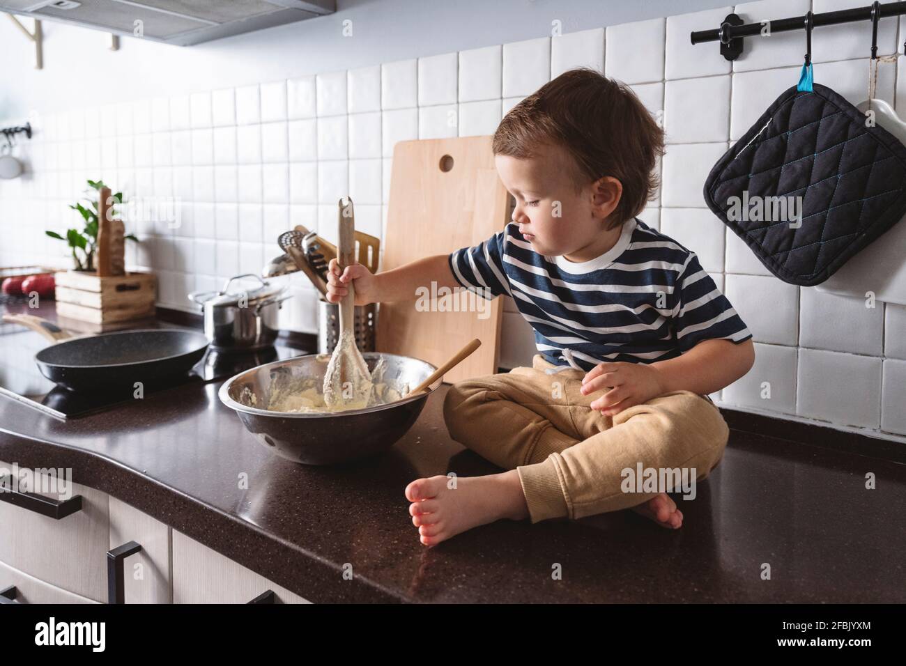 Netter Junge, der Zutaten in einer Schüssel mischt, während er in der Küche sitzt Zähler zu Hause Stockfoto
