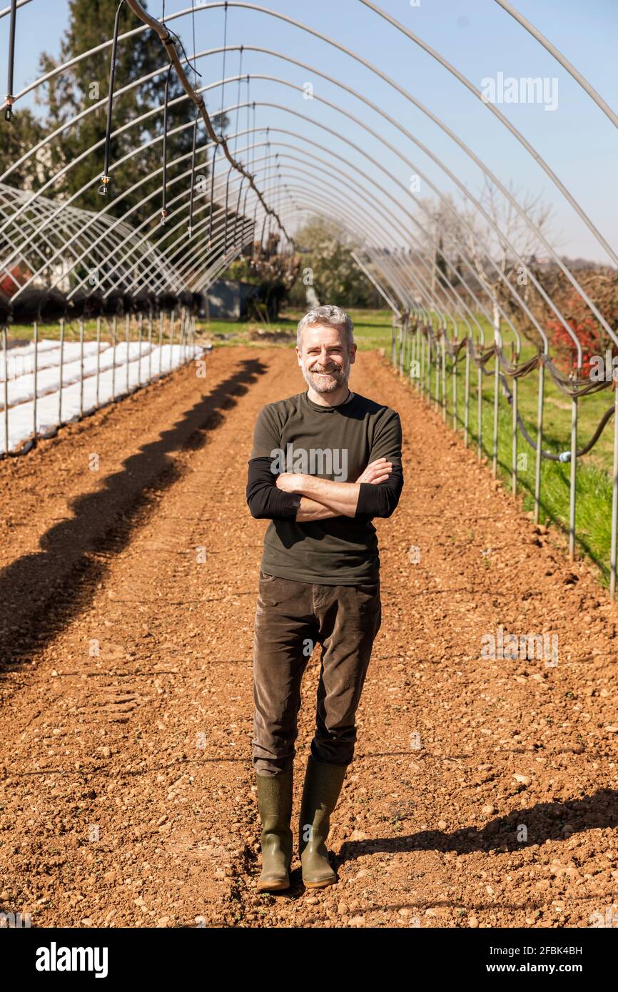 Lächelnder Bauer mit gekreuzten Armen, der auf dem Bio-Bauernhof steht Stockfoto