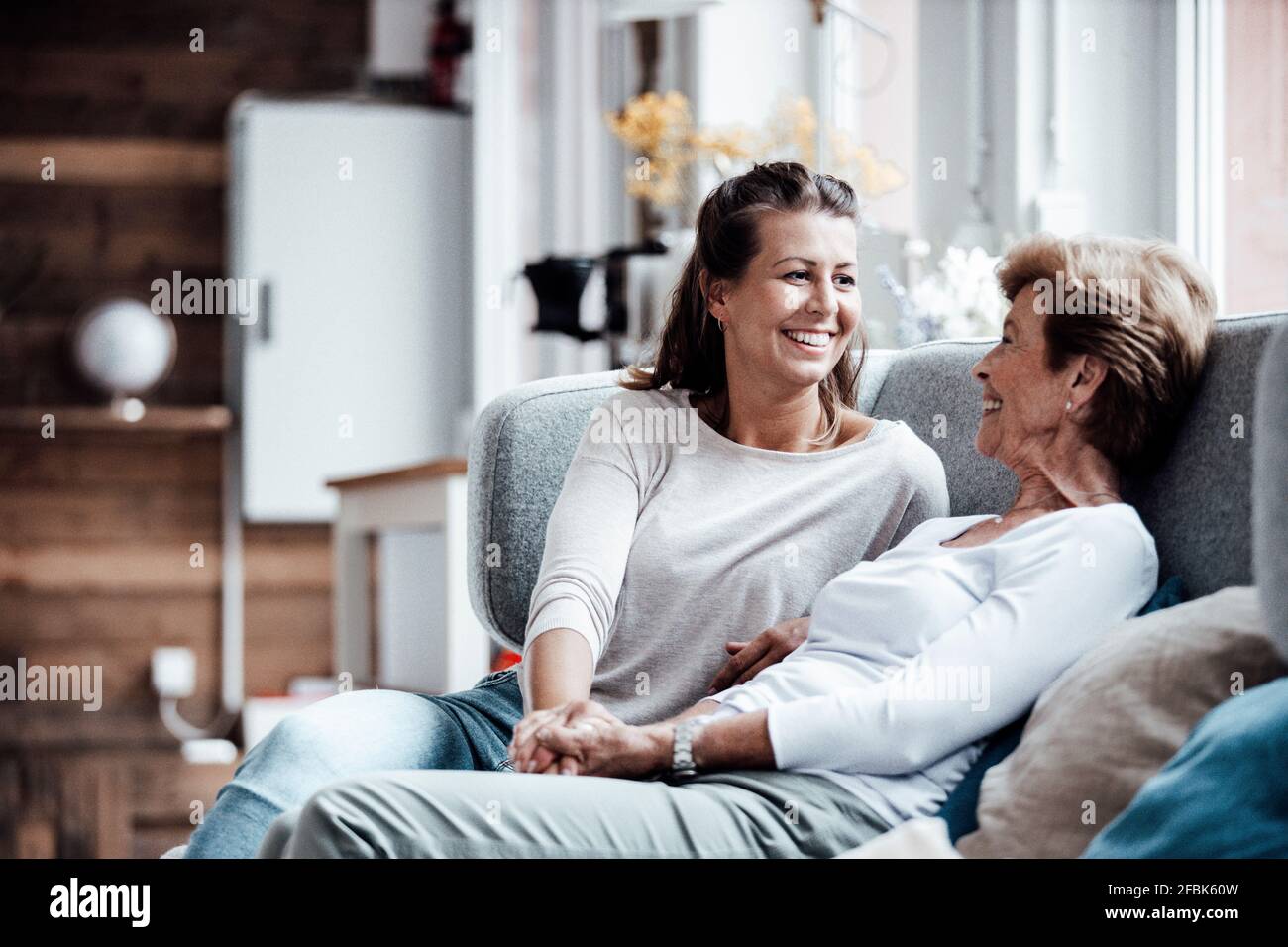 Lächelnde, schöne Frau, die die Hand der Großmutter hält, während sie auf dem Sofa sitzt Ein Zuhause Stockfoto