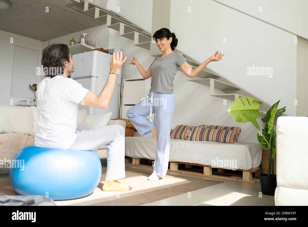 Frau, die dem Mann, der auf dem Fitnessball sitzt, Yoga beibringt Im Wohnzimmer zu Hause Stockfoto