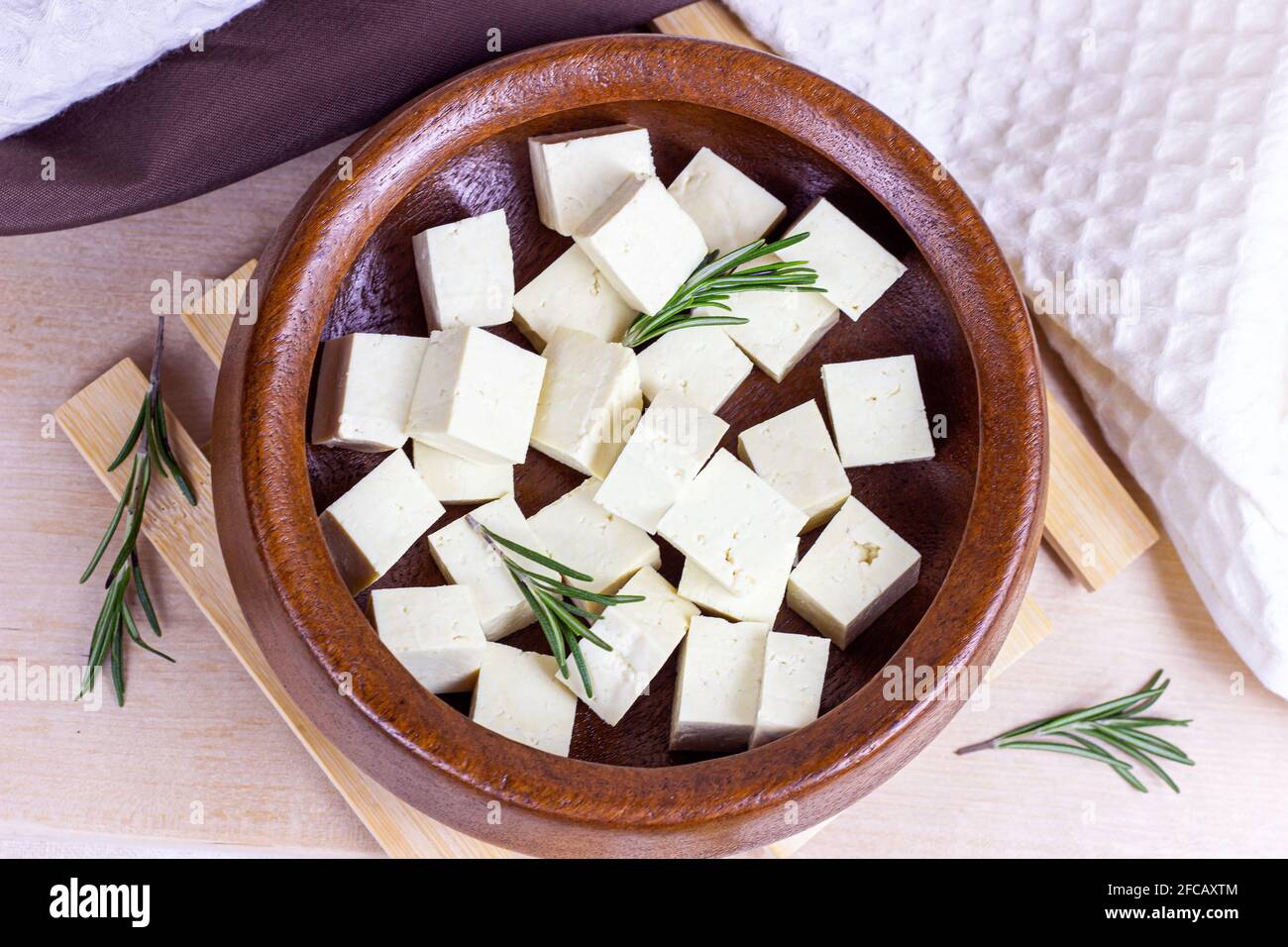 Draufsicht auf rohe Bio-Tofu-Würfel in runder Schüssel mit frischem Rosmarin auf Holzhintergrund. Stockfoto