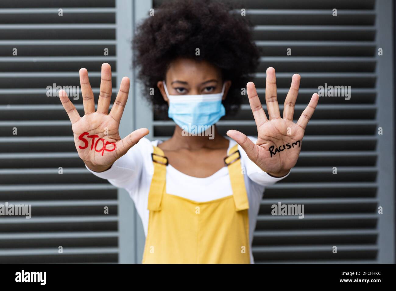 Portrait einer Frau mit gemischter Rasse, die Gesichtsmaske trägt, die Halt macht Geste mit schriftlich bedeckten Händen Stockfoto