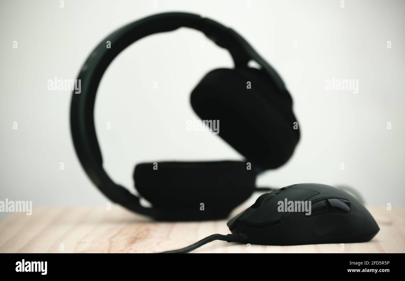 Kopfhörer und Maus in einem hölzernen Computertisch, Maus im Fokus als Headset verschwommen hinter, dunklere Objekte vor dem helleren Hintergrundfoto Stockfoto