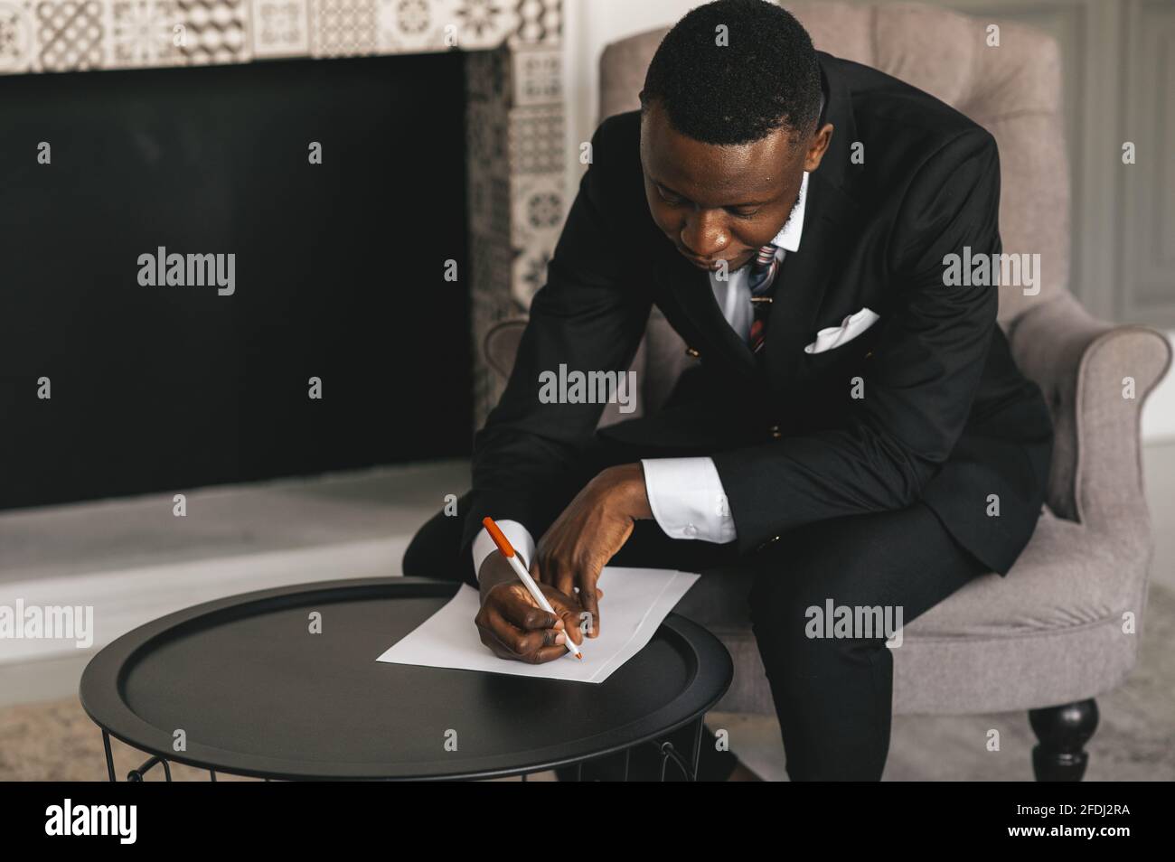 afroamerikanischer Geschäftsmann in einem Anzug macht Notizen auf einem Blatt Papier, während er in seinem Büro in der Nähe des sitzt Kamin Stockfoto