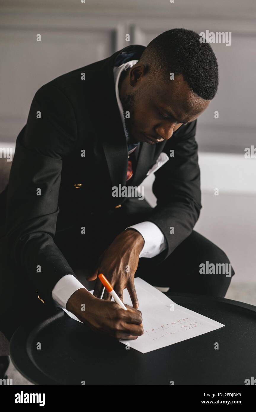 afroamerikanischer Geschäftsmann in einem Anzug macht Notizen auf einem Blatt Papier, während er in seinem Büro in der Nähe des sitzt Kamin Stockfoto