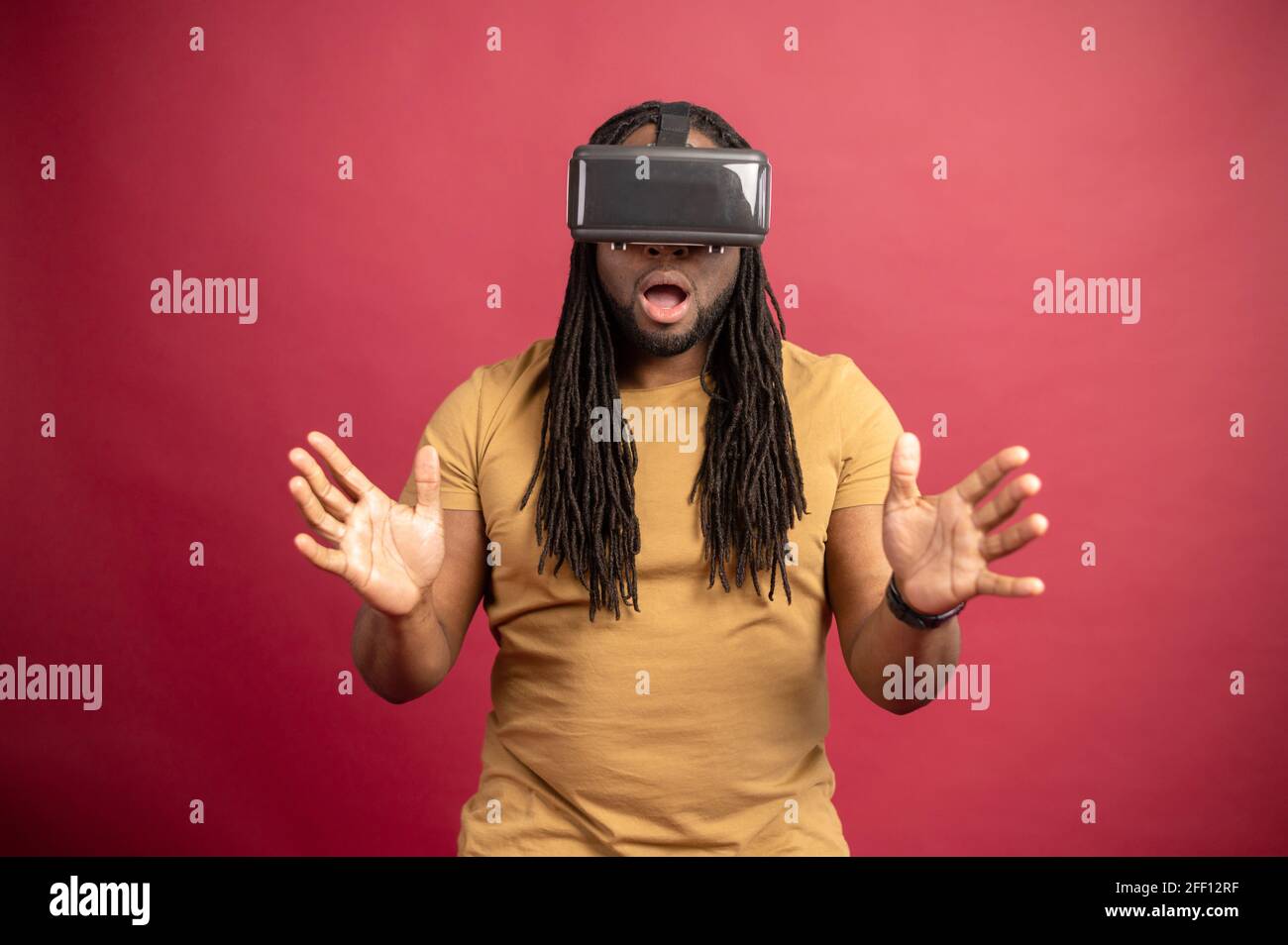 Überrascht verängstigten afroamerikanischen Mixed-Race-Mann mit Dreadlocks und offenen Mund isoliert über rote Wand berühren Luft mit Händen, verbringen Zeit in der virtuellen Realität, spielen Spiele in modernen VR-Headset Stockfoto