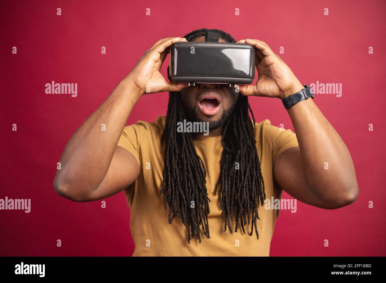 Schwarz staunender Mann mit langen Dreadlocks in legerer Kleidung, isoliert auf rotem Hintergrund stehend mit Virtual-Reality-Headset, Brille in einem leeren Raum, unterwegs im Internet, neue Erfahrung ausprobieren Stockfoto