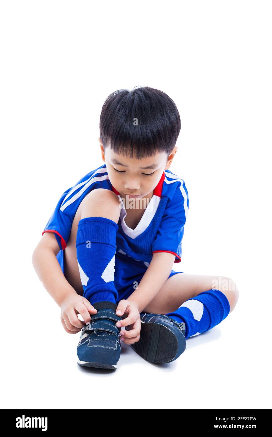 Junger asiatischer (thailändischer) Fußballspieler in blauer Uniform bindet Schuhe, bevor er Fußball mit Schlagschatten spielt. Kind bereitet sich auf den Wettkampf vor. Sportleben Stockfoto