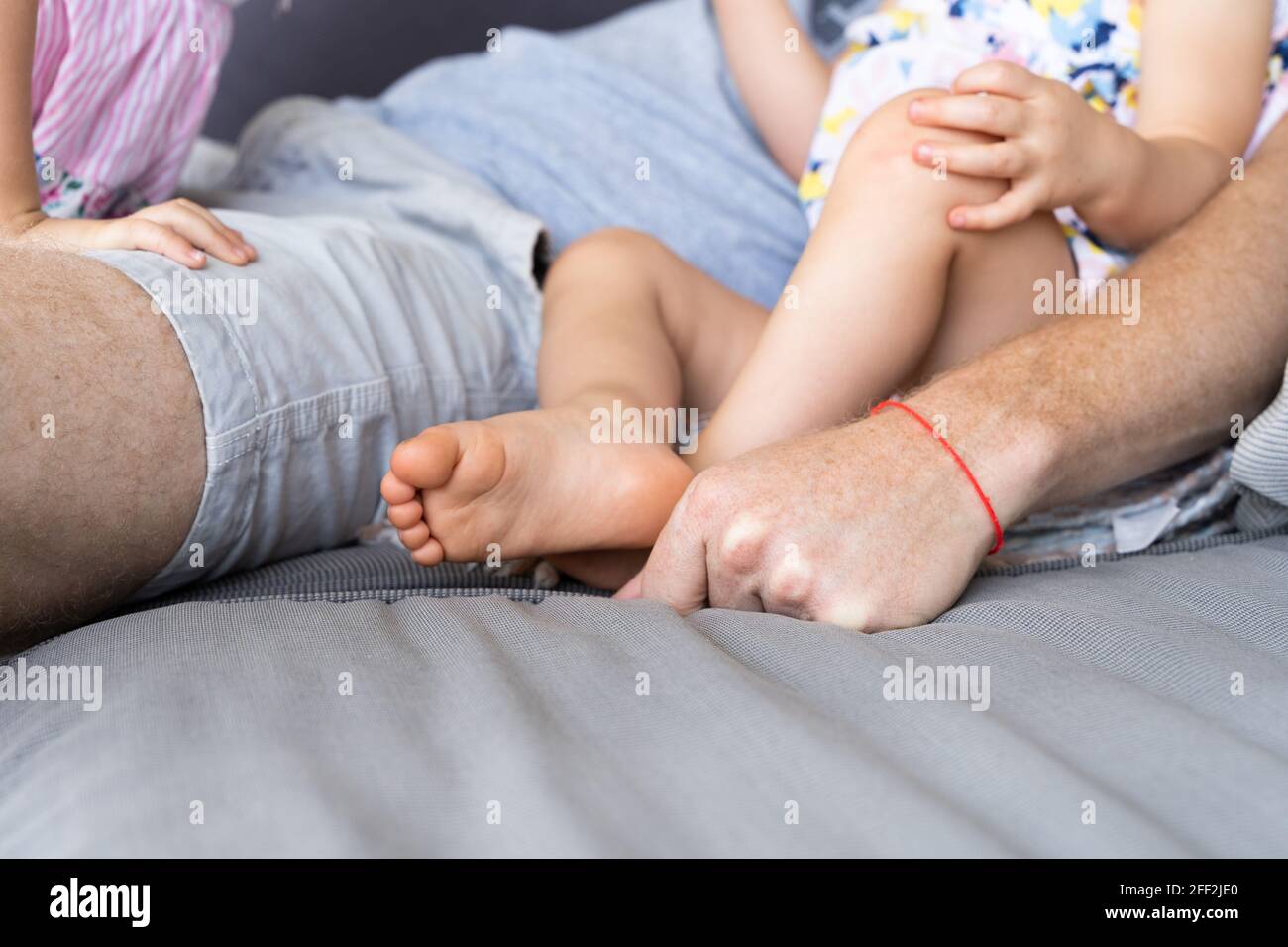 Anonymer Vater mit rotem Armband auf der Couch mit Baby-Mädchen. Vater sitzt mit kleinen Töchtern auf dem Sofa Stockfoto