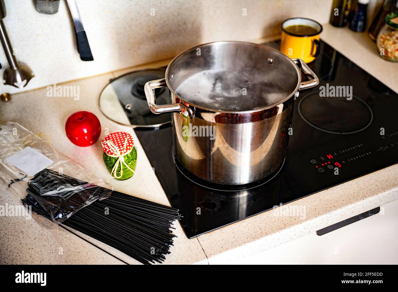Kochendes Wasser im Topf auf heissem Keramikkochfeld, Beutel mit schwarzen Spaghetti, Flasche mit grünem Basilikum-Pesto, Küchenuhr für roten Apfel, kleiner gelber Topf mit Suppe. Stockfoto