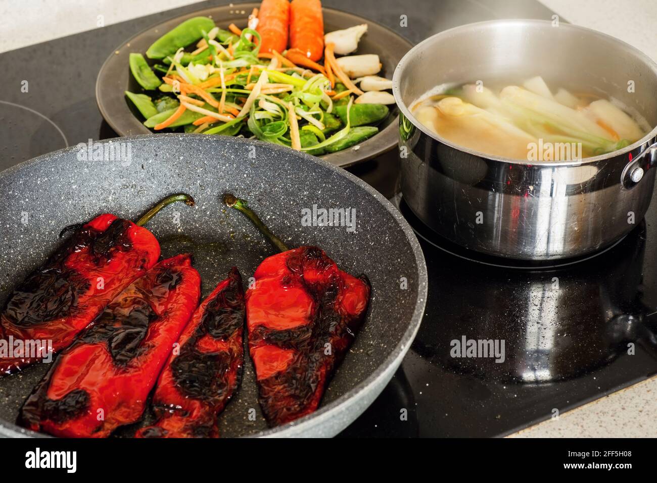Pfanne mit geröstetem Paprika, Topf mit kochendem Spargel und Teller mit geschnittenem Gemüse auf heissem Keramikherd. Vegetarische Gerichte werden zubereitet. Stockfoto