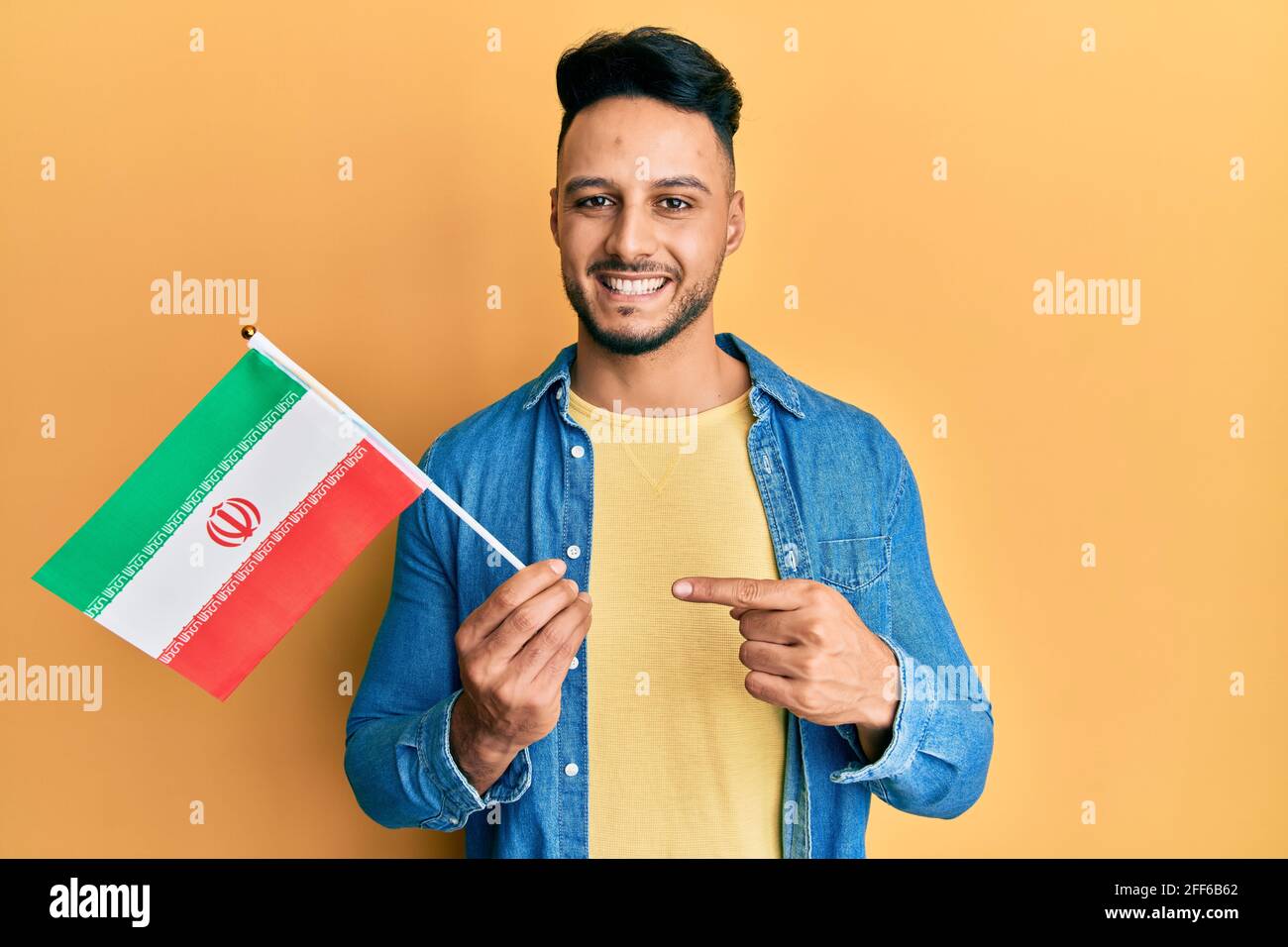 Junger arabischer Mann, der die iranische Flagge hält, lächelt glücklich und zeigt mit ihm Hand und Finger Stockfoto